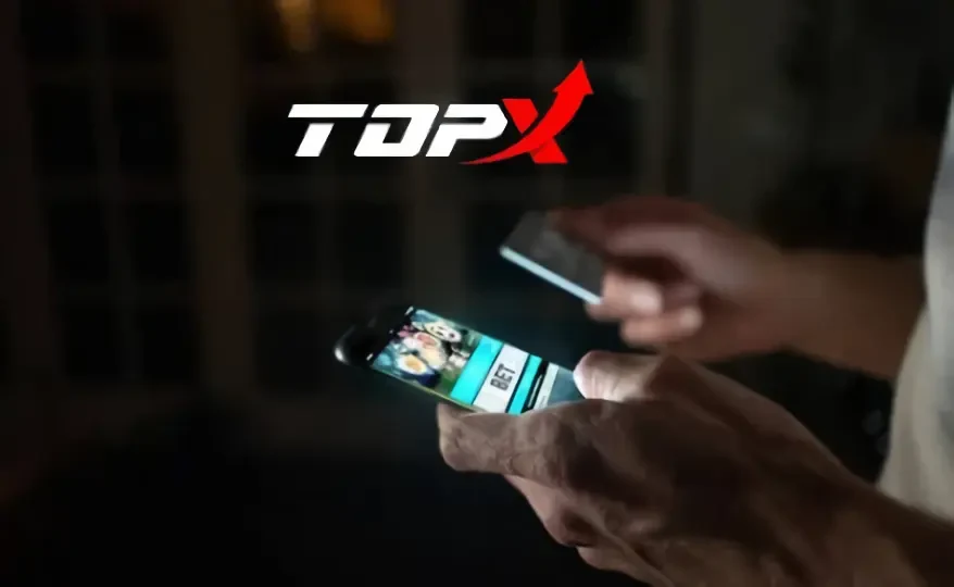 Mãos em ambiente escuro usando um celular com uma tela de cassino, com o logotipo da TopX acima